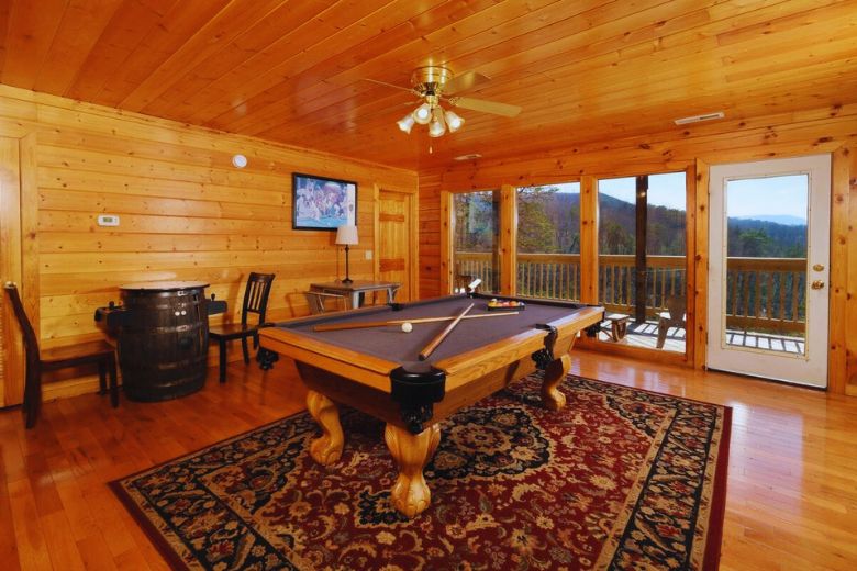 Cozy cabin game room with a pool table centered on a decorative rug, wood-paneled walls and ceiling, flat-screen TV, and large windows offering mountain views with access to a wraparound deck.